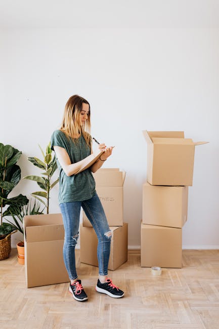 A young woman with shoulder-length blond hair, wearing a casual green t-shirt, ripped blue jeans, and black and pink sneakers, is standing in a well-lit room with a white wall and wooden flooring. She is holding a pen and a small notepad, appearing to take notes during a home relocation or packing process. Next to her, there are several cardboard moving boxes—some sealed and others open—stacked on the floor and against the wall, with one box prominently open at the top. Behind her, there is a large leafy potted plant placed on the floor, adding a touch of greenery to the space. The overall setting suggests preparation for a move, with her documenting packing details, as part of a furniture transport or home packing and moving service, possibly provided by Chelsea Man and Van, in the context of a Chelsea Harbour moving checklist for flats and penthouses.