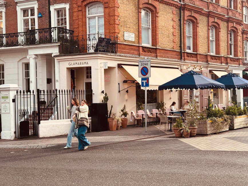The image shows the exterior of a brick-built building in Chelsea, with a ground-floor café featuring outdoor seating under large navy blue umbrellas. There are several tables with chairs, some occup
