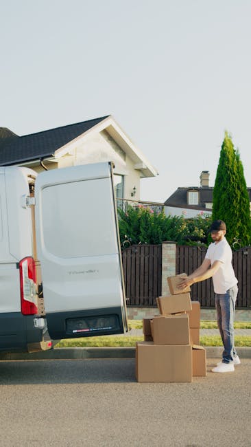A young man wearing a white t-shirt and jeans is standing outdoors on a residential street, preparing cardboard boxes for a home relocation. He is positioned next to an open white van, which is likely being used for furniture transport and packing and moving activities. The boxes are of various sizes, stacked on the pavement near the van, with some being lifted or ready for loading. The scene is illuminated by natural daylight, with a background featuring a modern house with a sloped roof, a wooden fence, and tall trees. The van is parked on the street, and the image captures the process of loading boxes into the vehicle, with visible packaging materials like boxes and potentially some plastic or fabric wrapping. This setting reflects typical moving logistics and the involvement of professional removal services, such as those offered by Chelsea Man and Van, as showcased on the Kings Road Chelsea moving guide page for removals.