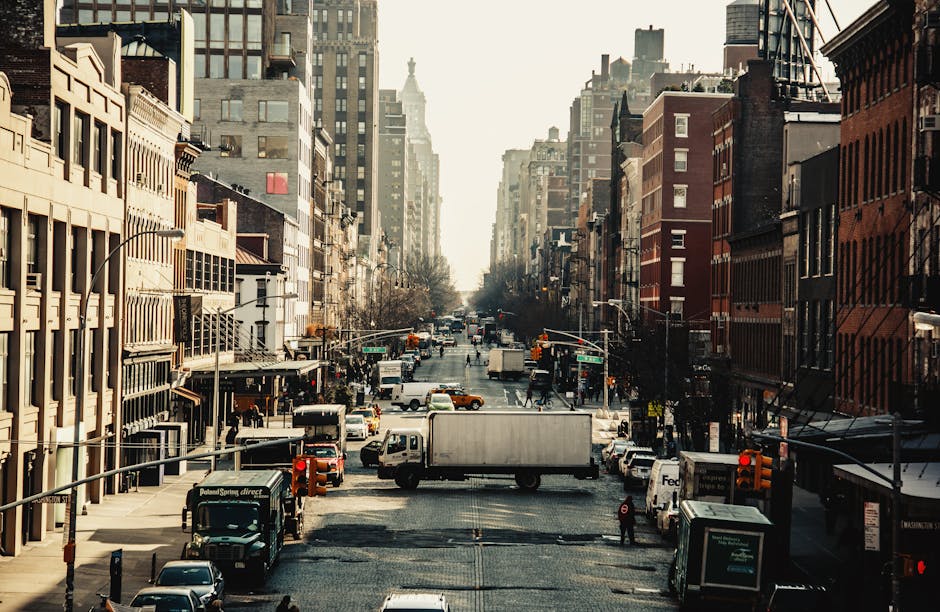 A busy city street scene in a metropolitan area with tall, multi-storey buildings lining both sides of the road, featuring a variety of commercial and residential structures with brick and concrete facades. The street is filled with parked and moving vehicles, including delivery vans, trucks, and cars, with some waiting at red traffic lights. The road has multiple lanes with traffic signals and pedestrian crossings visible, and a few pedestrians are present on the sidewalks. In the foreground, a large white delivery truck is parked on the street, possibly staged for a home relocation or furniture transport, with a few workers nearby handling packing materials such as cardboard boxes, plastic wrap, and moving blankets. Some boxes are stacked on dollies or trolleys, ready for loading or unloading, with furniture pieces partially wrapped in fabric or plastic. The scene is well-lit with natural daylight, capturing the logistical process involved in house removals and moving services, as operated by Chelsea Man and Van, in an urban environment consistent with the Kings Road Chelsea moving guide for man and van services.