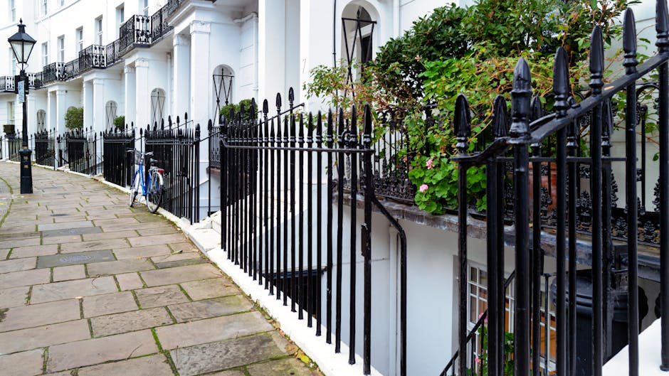 A row of white terraced houses with black wrought iron railings along the pavement on Sloane Square, with some greenery and potted plants visible on the stairs and window sills. A blue bicycle is leaning against the railing near an accessible basement entrance, which is open and surrounded by white stonework. The pavement is made of large stone slabs, and the scene is well-lit with natural daylight. This setting exemplifies typical narrow-access properties in Chelsea, where careful planning is required for home relocation and furniture transport, often involving packaging, loading, and unloading through confined spaces as managed by Chelsea Man and Van.