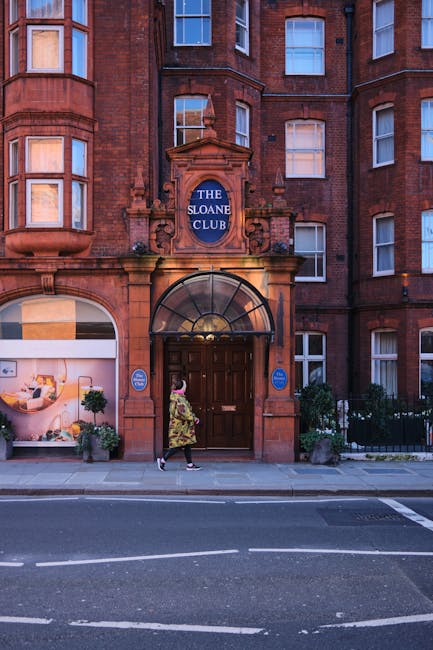 A woman wearing a floral dress and black shoes stands on the pavement outside a multi-storey red-brick building identified as 'The Sloane Club.' The building features a prominent entrance with a rounded glass canopy above dark wooden double doors, framed by stone detailing and flanked by two blue signs. The pavement is clear, with small potted plants near the entrance, and a large display window to the left showing a modern interior with furniture and decorative items. The scene depicts an urban setting suitable for house removals or furniture transport, with Chelsea Man and Van’s services connected to moving in or out of historic properties, and the lighting indicating late afternoon or early evening.
