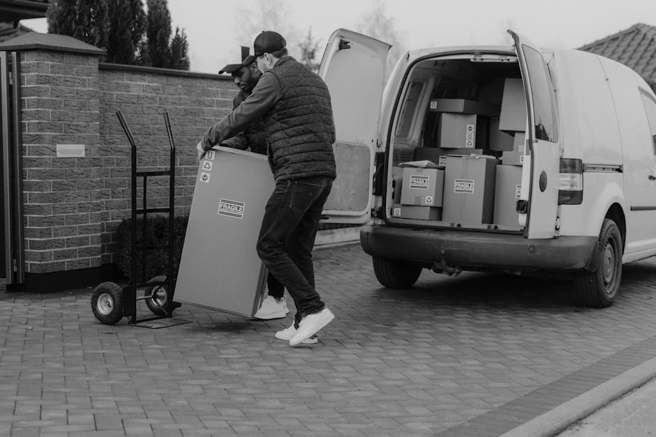 Two movers from Chelsea Man and Van are engaged in a home relocation process, loading a cardboard box onto a trolley outside a property. The van, positioned on a cobblestone driveway, has its rear doors open, revealing numerous packed boxes, some labelled with fragile symbols, indicating domestic packing and moving activities. One mover, wearing a cap and dark clothing, is assisting with lifting the box, while the other, dressed in a puffer jacket and jeans, supports it from the other side. The scene takes place outdoors near a brick wall, with the car positioned close to the entrance for easy transfer of household items. The environment appears overcast, with natural light illuminating the packing and loading movement, which is part of a furniture transport or moving logistics process managed by Chelsea Man and Van. This image captures the logistics of professional removals, including packing materials, equipment such as trolleys, and the loading process essential for successful house moves.