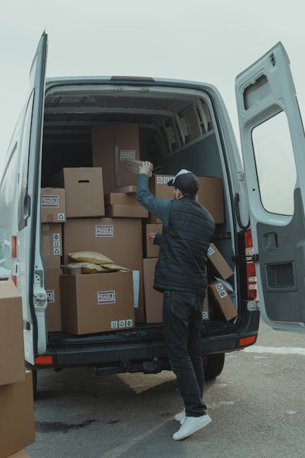 A person wearing a black cap, black jacket, and dark trousers is loading or unloading a variety of cardboard boxes into or from the back of a grey van during a furniture transport or home relocation process. The interior of the van is visible, showing additional boxes stacked neatly, some wrapped in packing materials such as bubble wrap or paper. The boxes are of different sizes, some labeled with small symbols or text, indicating their contents or handling instructions. The person is standing on a paved surface, using their right hand to lift or position a box, while other boxes remain inside the vehicle and on the ground nearby. The scene takes place outdoors with a clear sky, and the van appears to be part of a professional removals service, such as Chelsea Man and Van, engaged in a packing and moving operation. The open rear doors of the van reveal a spacious interior designed for efficient furniture and box loading, supporting quick and organized relocation services, including same-day or quick removals commonly provided by companies like Chelsea Man and Van.