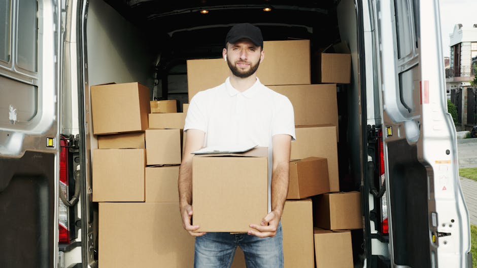 Two movers from Chelsea Man and Van are engaged in a home relocation process, loading a cardboard box onto a trolley outside a property. The van, positioned on a cobblestone driveway, has its rear doors open, revealing numerous packed boxes, some labelled with fragile symbols, indicating domestic packing and moving activities. One mover, wearing a cap and dark clothing, is assisting with lifting the box, while the other, dressed in a puffer jacket and jeans, supports it from the other side. The scene takes place outdoors near a brick wall, with the car positioned close to the entrance for easy transfer of household items. The environment appears overcast, with natural light illuminating the packing and loading movement, which is part of a furniture transport or moving logistics process managed by Chelsea Man and Van. This image captures the logistics of professional removals, including packing materials, equipment such as trolleys, and the loading process essential for successful house moves.
